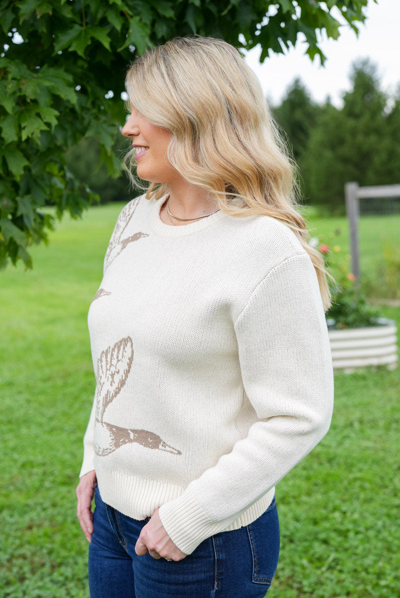 A woman wearing a cream-colored knit sweater with brown mallard duck illustrations, standing outdoors in a grassy field with trees in the background.
