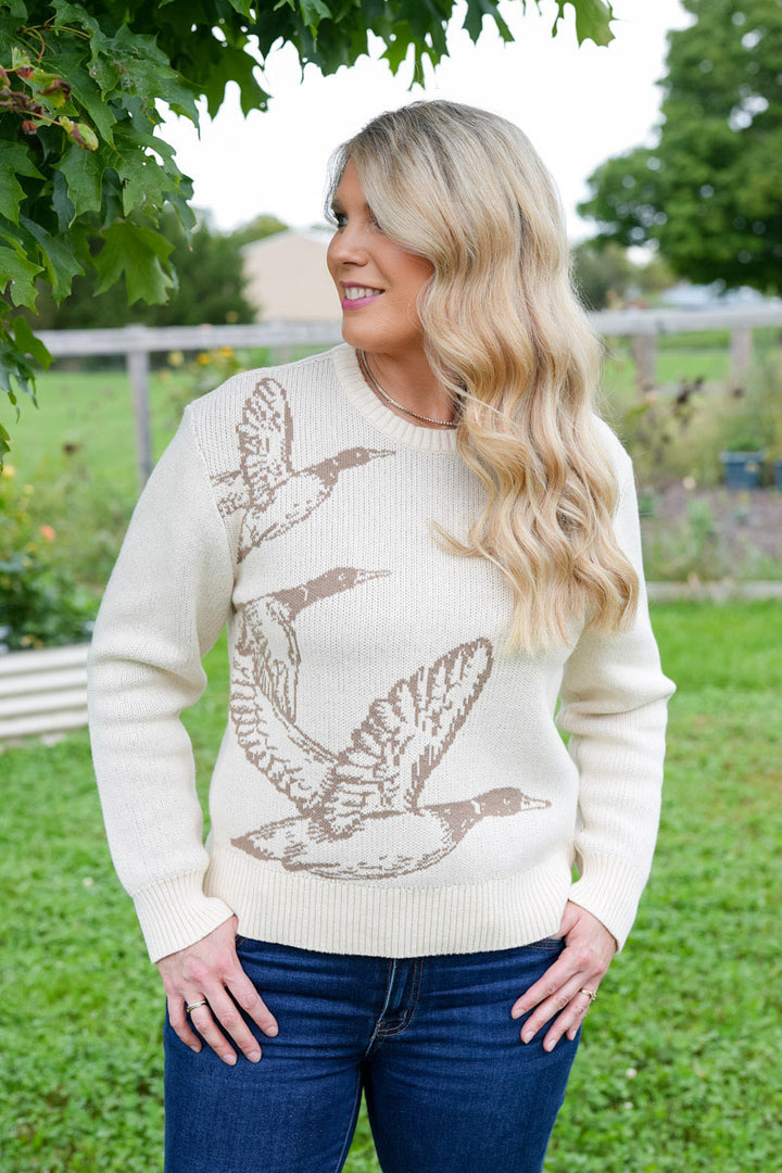 A woman wearing a cream-colored knit sweater with a brown mallard duck hunting bird graphic, paired with blue jeans, standing outdoors in a grassy yard.