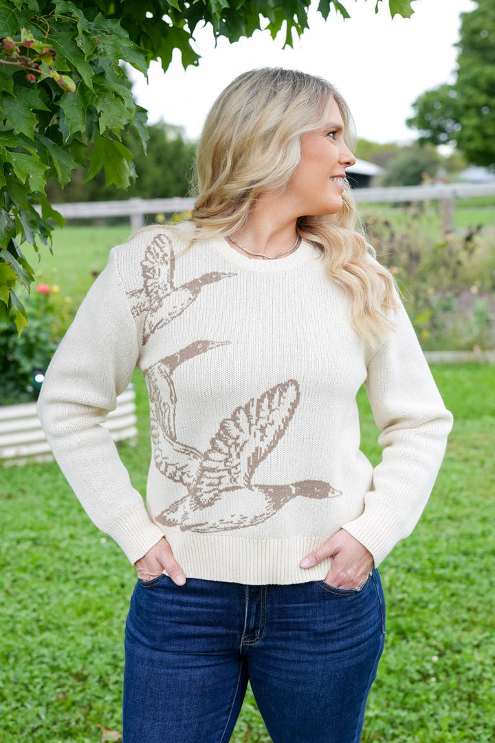 A woman wearing a cream-colored knit sweater with a brown mallard duck hunting bird pattern, paired with dark blue jeans, standing outdoors in a grassy field.