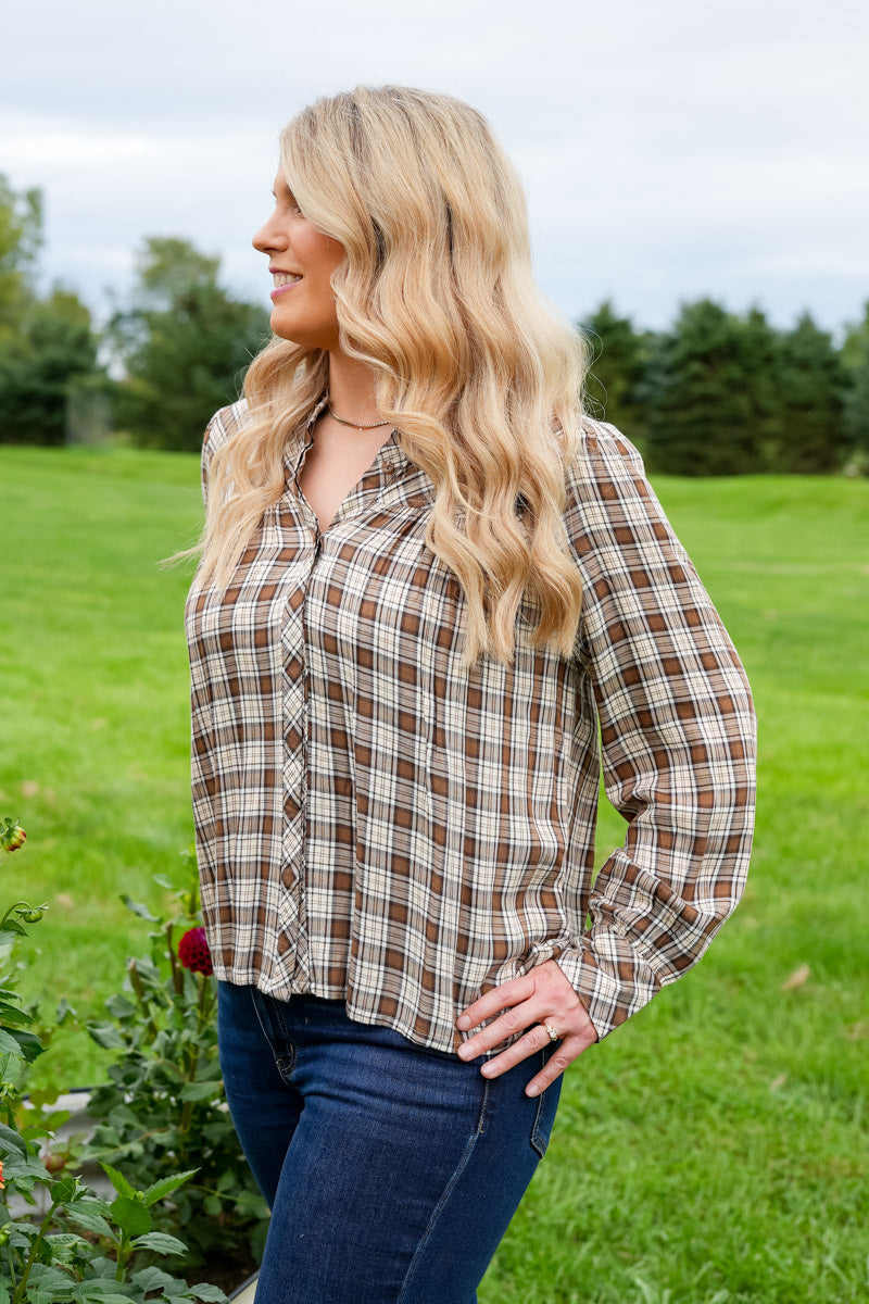 A woman with long blonde hair stands outdoors in a grassy garden wearing a brown and beige plaid button-down shirt and dark blue jeans.