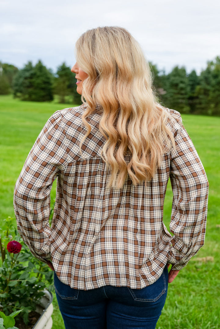 A woman with long blonde wavy hair is shown from behind wearing a brown and beige plaid button-up shirt and blue jeans, standing in a grassy outdoor field.