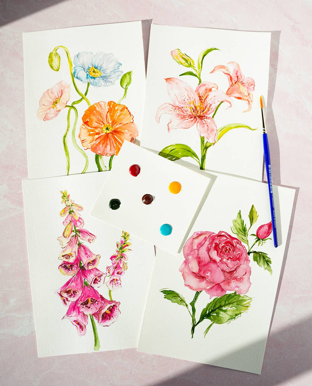 Four floral paintings on white paper with a paintbrush and paint dots on a light pink background.