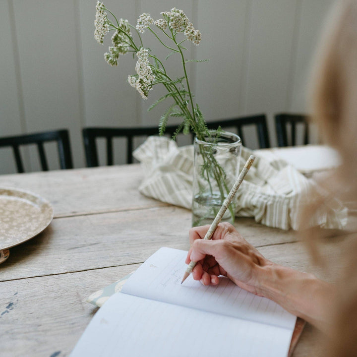 Person writing in a notebook with a vase of flowers on a wooden table