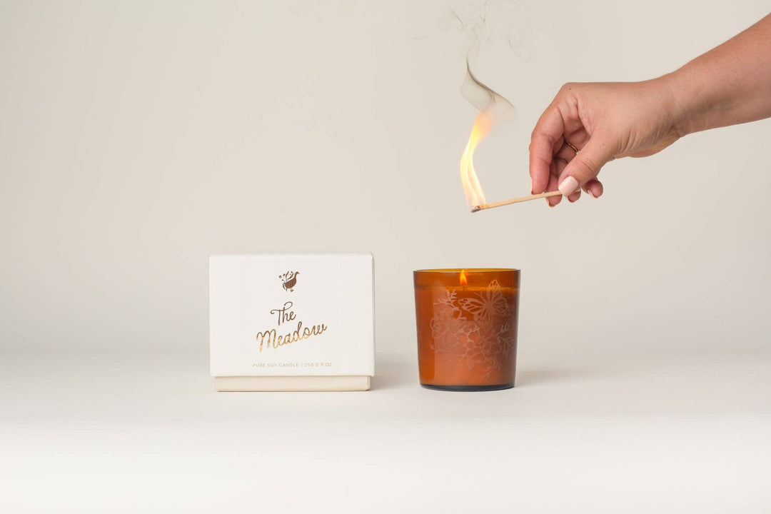 Hand lighting a candle next to a box labeled 'The Meadow' on a light background