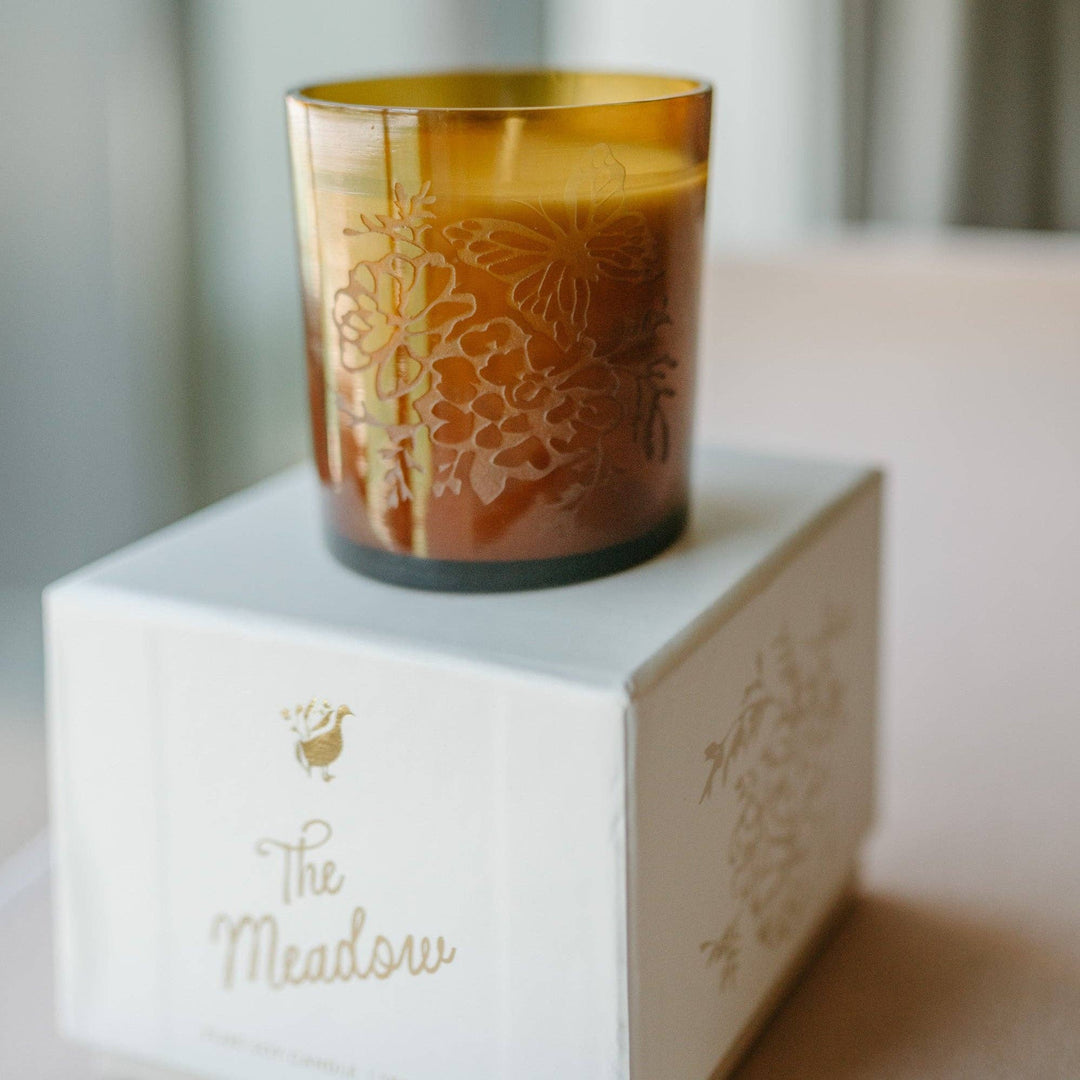 Decorative candle in a brown glass holder on a white box with 'The Meadow' branding.